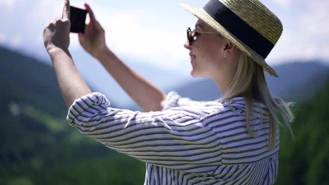Young caucasian woman in trendy hat making picture on modern mobile phone camera explore scenic landscape of Apls, breathtaking nature of mountains, female blogger photographing nature at journey