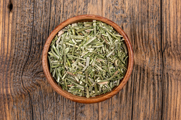 A wooden bowl filled with whole dried rosmary on a rustic wooden background