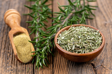 A wooden bowl filled with whole dried rosmary, a spice shovel with ground rosemary and two rosemary...