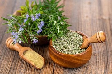 A wooden bowl with blooming and fresh rosemary twigs, a wooden bowl with whole dried rosmary and a spice shovel with ground rosemary on a rustic wooden background