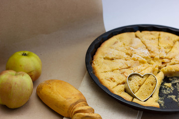 apple pie with a heart shape on a linen tablecloth on a table with parchment paper next to a red-green apples and a wooden spoon with a vintage handle
