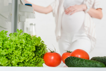 Selective focus of pregnant woman opening fridge with fresh vegetables isolated on white