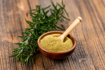 A small wooden bowl with ground rosemary and a small spice shovel and fresh rosemary twigs on a rustic wooden background