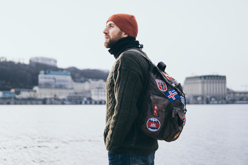 Young bearded traveller with patches on backpack standing at the river front with cityscape on...