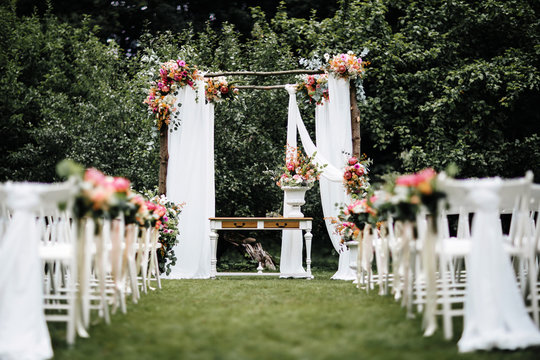 Decorated Luxury Wedding Ceremony Place In The Garden. White Empty Chairs And Arch Decorated With Flowers.