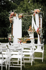 Decorated luxury wedding ceremony place in the garden. White empty chairs and arch decorated with flowers.