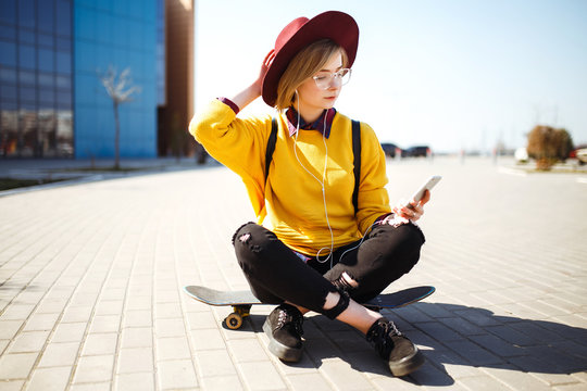 Young teenage girl sitting on a skateboard and texting on a mobile phone. Hipster girl in a yellow sweater, hat and sunglasses drinking from glass, with phone in her hands posing on skateboard. 