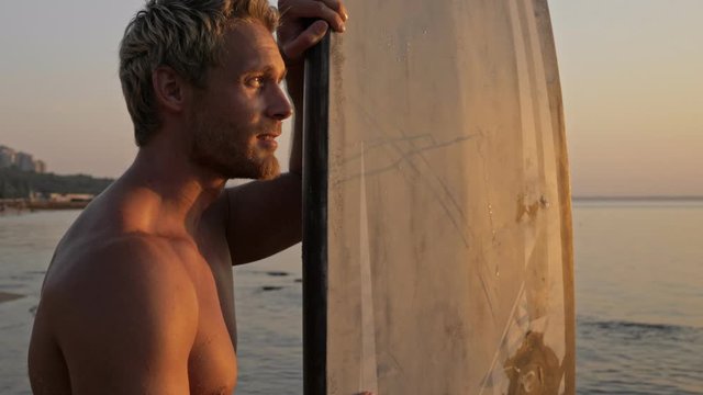 Smiling bearded surfer standing with surfboard and looking away on sunset near the sea