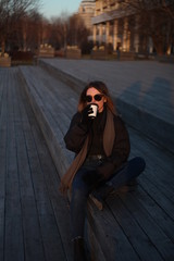 Fototapeta premium Young beautiful girl in sunglasses and a cap sits on the street and drinks coffee
