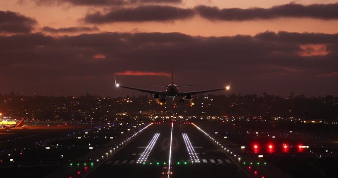 Impressive view of big modern airport at dark with lots of colorful lights all over it. Landing stripe is brightly illuminated. Landing plane appears from above, it descends and touches the ground. 4K