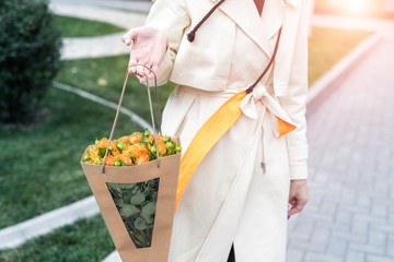 Close-up young adult fashion classy woman model hand holding authentic fresh orange spray roses flower bouquet wrapped in craft paper bag on city urban street outdoor. Spring present gift dilevery