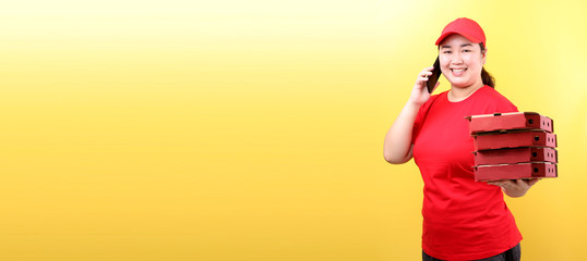 Asian Woman in red cap, giving food order italian pizza in cardboard  boxes isolated on yellow background in studio With copy space. holding mobile phone with blank  white  empty screen. 