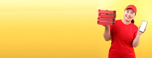 Asian Woman in red cap, giving food order italian pizza in cardboard  boxes isolated on yellow background in studio With copy space. holding mobile phone with blank  white  empty screen. 