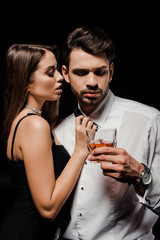 seductive woman touching bow tie of handsome man with glass of whiskey isolated on black
