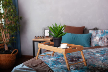 Bedroom interior with a small table on bed and a cup of coffee. Wooden breakfast tray on bed in morning. A cup of espresso in morning to bed. Honeymoon, surprise. Aloe Vera in a pot table. Room decor