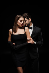 man in formal wear hugging beautiful woman in gloves isolated on black