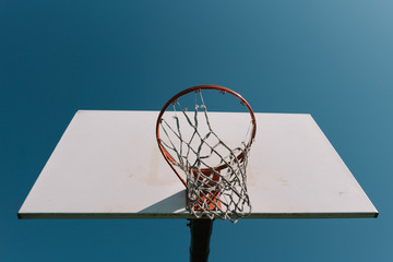 Looking up at old basketball hoop with torn net and white backboard alone against blue sky.