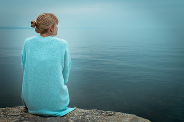 Beautiful single solo young traveler blond woman with tranquit jacket sitting alone on stone looking in to water in the blue ocean foggy mist lake in cold winter with sad dark depression  emotional - Powered by Adobe