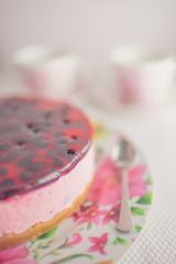 Part of round currant cheesecake in floral dish with spoon on a white table. On a blurred background two tea cups.