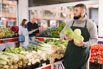 Male shop assistant lays fresh cabbage on counter in grocery shop