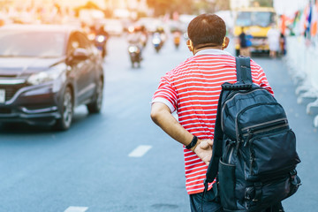 Fototapeta premium Back view of male patient with mask in red and white shirt with a black backpack standing at bus stop and waving his hand for Taxi or Bus in the city to go to the hospital.