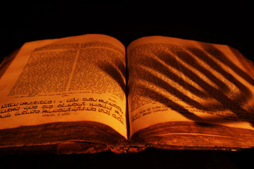 Old Hebrew Bible in light of burning candle on dark background. Shadow from menorah on open Jewish...