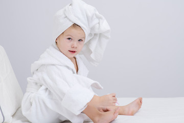 Little cute girl sitting on the bed in a white bathrobe and a towel on her head. Nail polish on toes. Little girl doing herself a pedicure