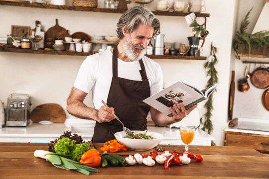 Image of happy gray-haired man in apron smiling and preparing recipe