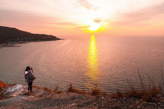 A woman with a backpack is standing on the edge of a cliff and taking photos of the sunset on her smartphone. In the background, the sea and the sky. Copy. Concept of Hiking and outdoor activities