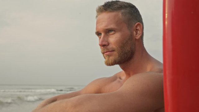 Smiling bearded handsome man looking away while sitting with surfboard on beach near the sea