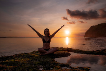 Excited young woman sitting on the beach, raising arms in front of the ocean. Sunset golden hour at the beach. Bali, Indonesia.