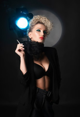 A beautiful blonde girl with an elegant hairstyle and large breasts, wearing a bra, trousers, scarf and a blazer, smokes a cigarette near the spotlight. Trendy, commercial and advertising design.