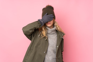 Young girl with winter hat isolated on pink background covering eyes by hands