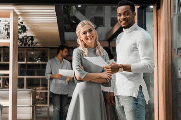 smiling african american businessman holding documents while standing near attractive colleague in office