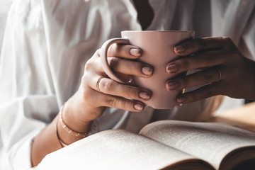 Woman in the morning drinks coffee and reads old book in a white shirt. Education, drink. manicure
