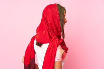 Young Indian woman with colorful holi powders on her face isolated on pink background in back position and looking side