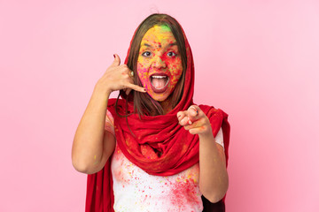 Young Indian woman with colorful holi powders on her face isolated on pink background making phone gesture and pointing front