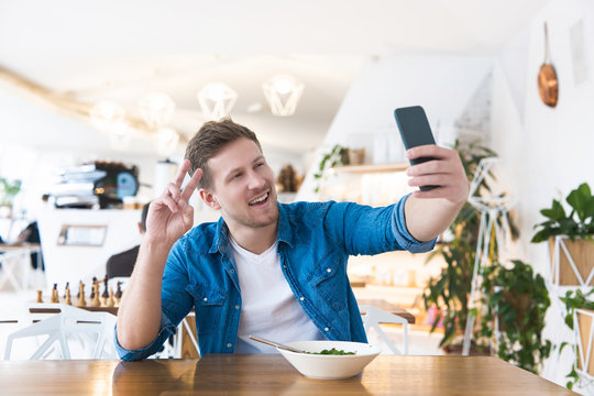 young handsome smiling man speaking online in his smartphone while eating salad during his lunch break in the cafe , multitasking concept