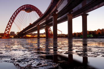The photo shows picturesque bridge with big red arch over the river. This cable-stayed bridge stands on the frozen Moscow river. Crimson sunny rays illuminate bridge details and ice on the sunset. 