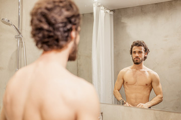 young caucasian man examining his face in mirror. half-naked guy with muscular body stand in bathroom after shower. hygiene, people concept