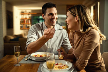 Happy man feeding his girlfriend while having dinner in dining room.