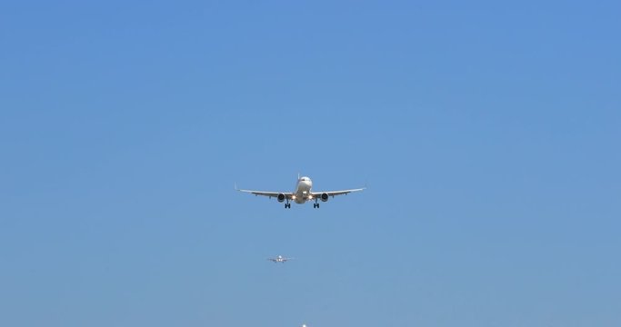 Frontal shot of modern white airplane landing, with landing gears seen released. Clear blue sky is all around the plane. Airliner smoothly descends approaching the camera and flies right above it. 4K