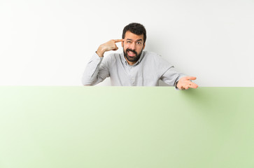 Young handsome man with beard holding a big green empty placard making the gesture of madness putting finger on the head