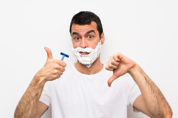 Man shaving his beard over isolated white background making good-bad sign. Undecided between yes or not