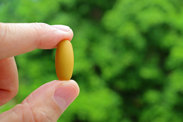 Man's Fingers Holding a Pill with Blurry Green Foliage in Background
