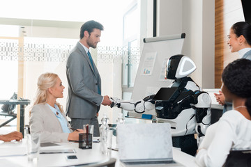 handsome businessman shaking hands with robot near multicultural colleagues sitting in conference hall