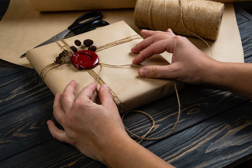 Obraz na płótnie Canvas Process of package gift wrapped in craft paper, tied with string and glued wax seal on wooden black background