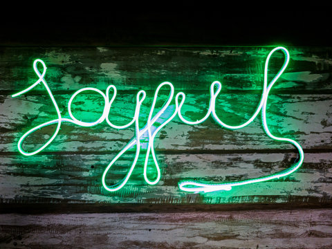 Green neon signage word Joyful made of LED strip light green color on old wood plank backdrop, dark theme background photography.