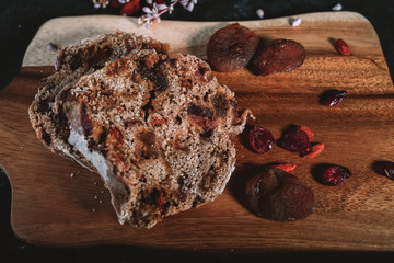 Homemade fruit bread on wooden chopping board with scattered dry berries and flowers on background on black table.