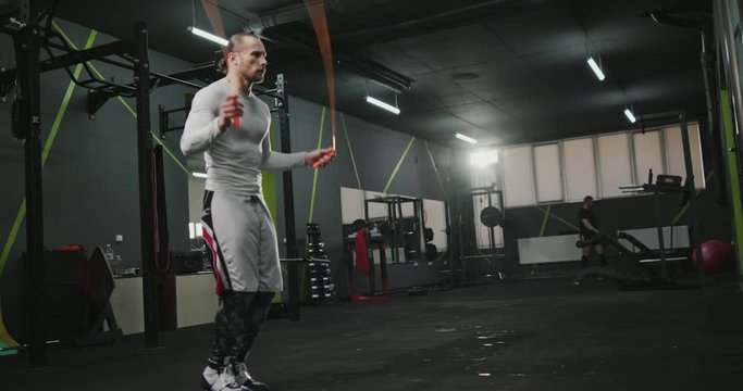 Sportive muscular man in grey training sportswear working out with skipping rope in gym. Low angle footage with copy space